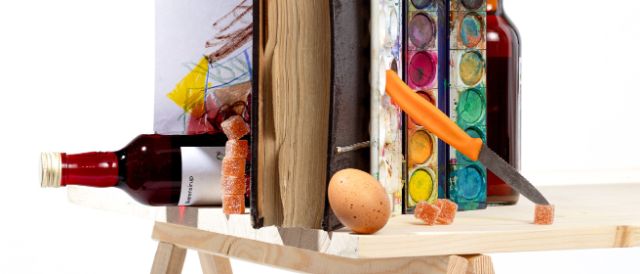 Die Fotografie ist als Stilleben arrangiert und zeigt auf einem Holztisch Bücher, Farbkasten, Sirupflaschen, ein Ei, Würfelzucker und ein Messer mit orangem Griff. Kinderzeichnung und Malspuren deuten auf kreative Arbeit hin. Die Objekte sind ungewöhnlich kombiniert und kunstvoll arrangiert. Sie scheinen die Schwerkraft zu umgehen.